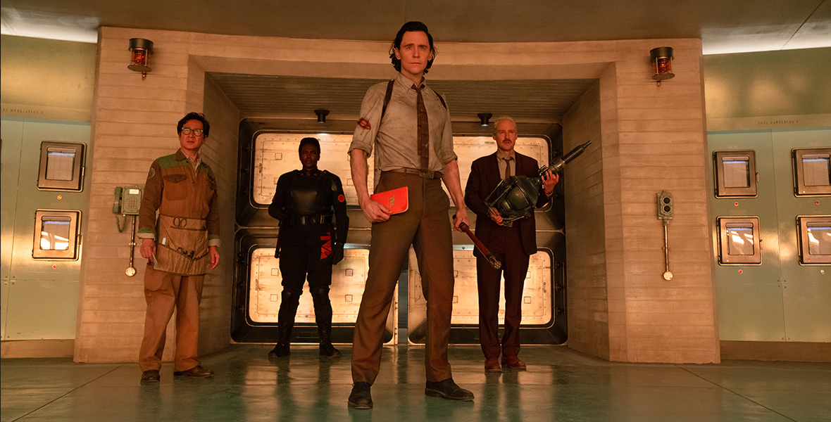 O.B., played by Ke Huy Quan, Hunter B-15, played by Wunmi Mosaku, Loki, played by Tom Hiddleston, and Mobius, played by Owen Wilson, stand inside the retro Time Variance Authority headquarters. Each of them is illuminated by an orange light.