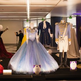 A wide shot of costumes from live-action Disney films focused on royalty with Cinderella’s iconic blue dress and Prince Charming’s ballroom look on mannequins at the center. At right is the red strapless gown worn by Anne Hathaway in Princess Diary. At left, a gold-toned outfit worn by Julie Andrews in the same film.