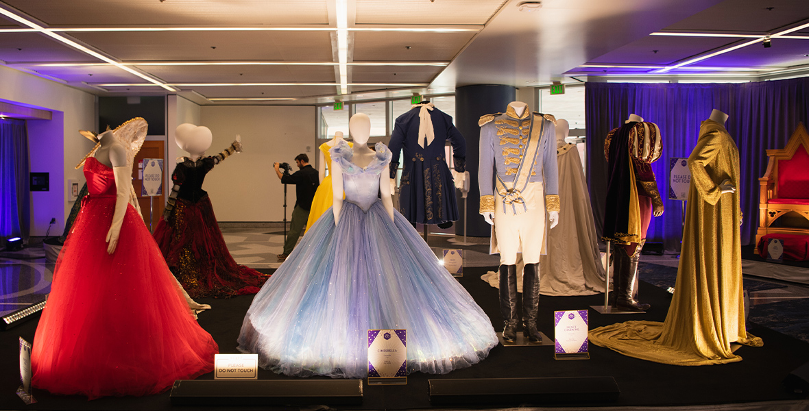 A wide shot of costumes from live-action Disney films focused on royalty with Cinderella’s iconic blue dress and Prince Charming’s ballroom look on mannequins at the center. At right is the red strapless gown worn by Anne Hathaway in Princess Diary. At left, a gold-toned outfit worn by Julie Andrews in the same film.