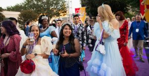 Guests walk on the purple carpet with red and orange banners behind them featuring crowns. Some guests have their phones up to film and others look at the people around them. They are all dressed up in gowns and suits.