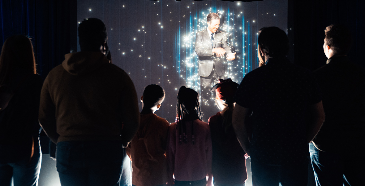 Guests are seen from the back in silhouette in the entrance room of Disney100: The Exhibition, as they watch a projected image of Walt Disney brushing pixie dust off of his arms on the Disney MagicStage during his opening greeting message.