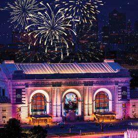 Fireworks light up the night sky behind Union Station in Kansas City, Missouri. The building is lit with yellow light and features banners for Disney100: The Exhibition hung over the entrance.