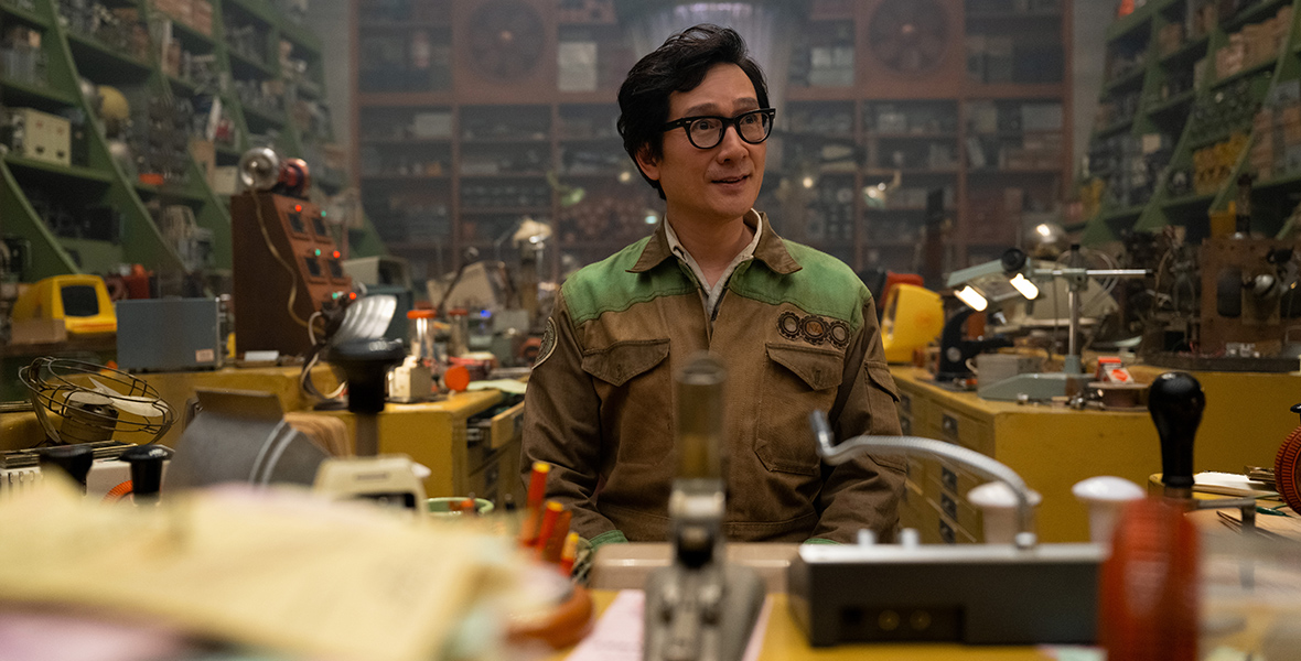 O.B. (Ke Huy Quan) sits at a messy desk in front of a complicated array of shelving inside the TVA. He is wearing a tan and grey boiler suit with three round patches on the right side of his chest.