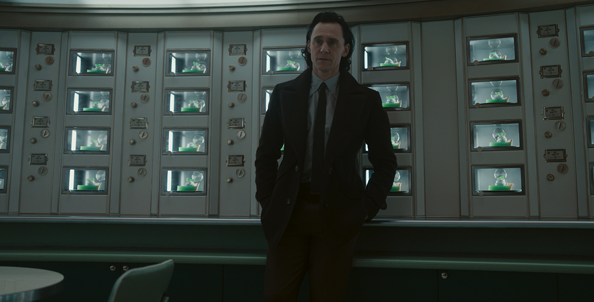 DAM1140_204_comp_INH_v0001.1053_R ALT TEXT: Loki (Tom Hiddleston) wears a suit and leans against a wall of cases, all holding bright green slices of key lime pie.