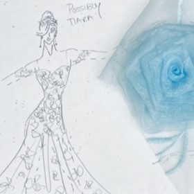 A sketch shows a woman wearing a dress that has a fitted bodice and a flowy skirt. It has roses all over it and a blue rose made of fabric is paper clipped to the paper. The woman in the sketch is also wearing a tiara.