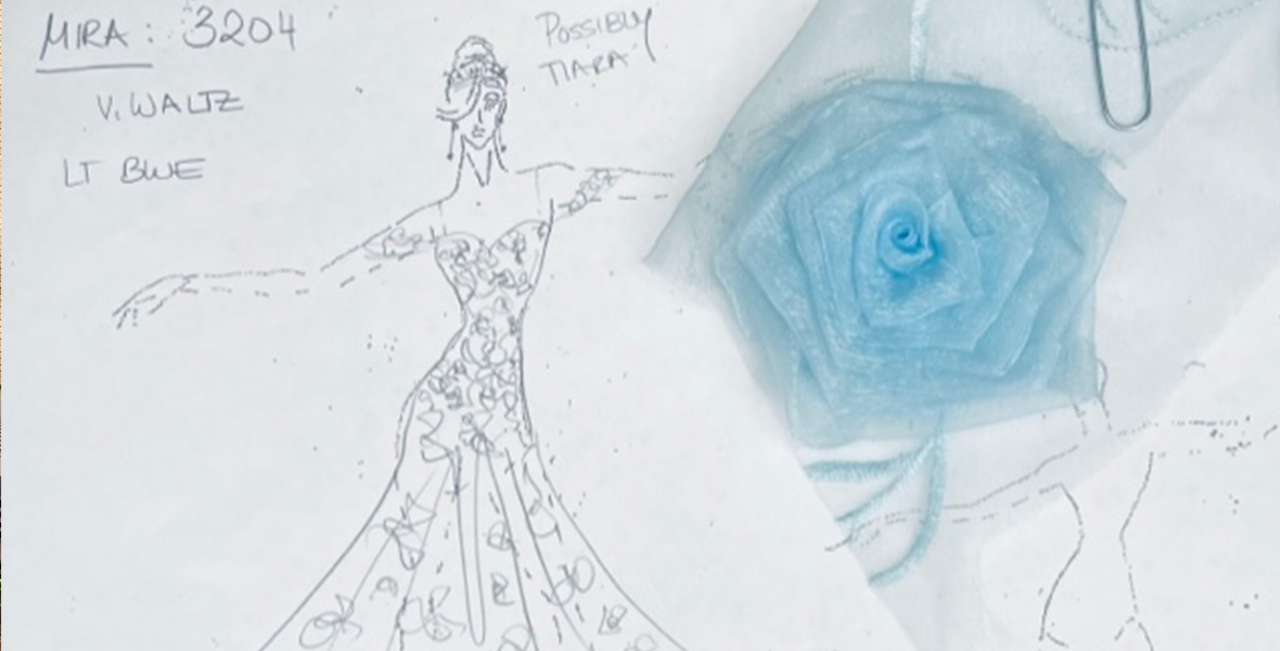 A sketch shows a woman wearing a dress that has a fitted bodice and a flowy skirt. It has roses all over it and a blue rose made of fabric is paper clipped to the paper. The woman in the sketch is also wearing a tiara.