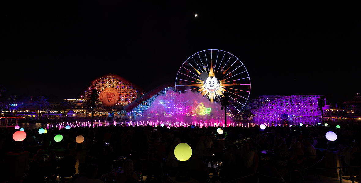 A picture of Disney California Adventure at night shows a Ferris wheel with Mickey Mouse’s face and the Incredicoaster attraction lit up with purple and orange lights.