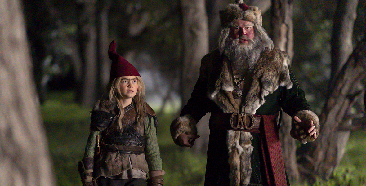 In an image from The Santa Clauses Season 2,  Magnus Antas (Eric Stonestreet) is dressed in a brown coat with a fur rim, gray pants, a fur hat, and brown boots; he has a grey beard and long hair. He’s standing next to a boy wearing an elf hat, green top, and black pants. The pair is standing in a forest.