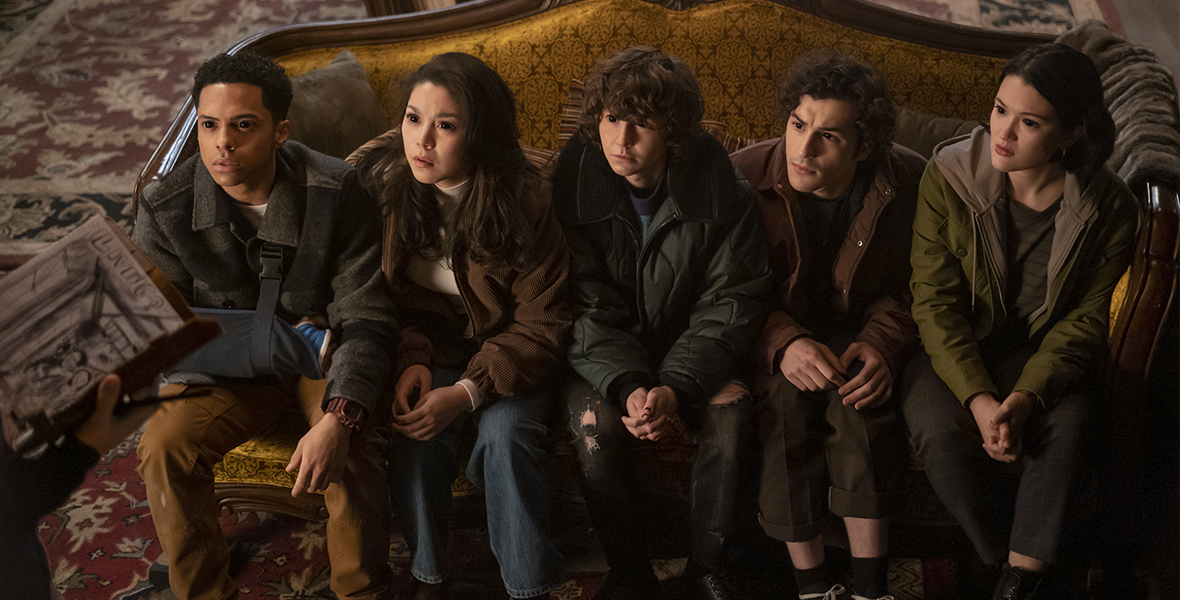 The 5 main characters from the new series “Goosebumps” sit on a yellow couch with a wooden frame while looking up at someone out of frame who is holding a book. The eyes of all five are fully black, with no irises or white areas visible. From left to right, they are Isaiah played by Zack Morris, wearing a gray coat and brown pants; Isabella played by Ana Yi Puig, wearing a brown coat and blue jeans; James played by Miles McKenna, wearing a green puffer coat and ripped black jeans; Lucas played by Will Price, wearing a brown jack and black jeans; and Margot played by Isa Briones, wearing a green jacket with a gray hoodie and black jeans.