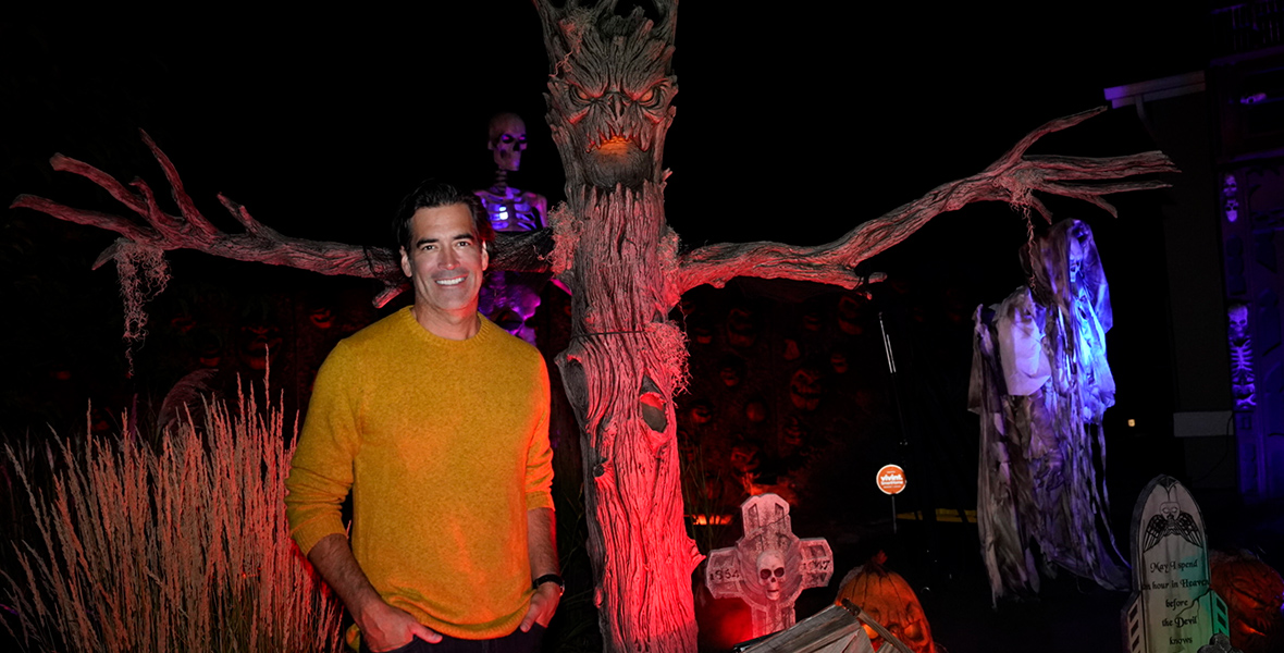 In a promotional image for ABC’s The Great Halloween Fright Fight, judge Carter Oosterhouse stands in front of one of the episode’s homemade Halloween displays. He’s wearing a goldenrod yellow sweater; to his right is a spooky tree with “arms” outstretched and a creepy face, surrounded by some tombstones.