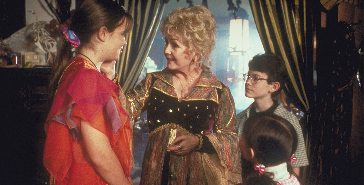 In an image from the 1998 Disney Channel Original Movie Halloweentown, Marnie (Kimberly J. Brown), left, is speaking with her grandmother Aggie (Debbie Reynolds), middle, while her siblings Dylan (Joey Zimmerman) and Sophie (Emily Roeske), right, look on. The family is standing in a living room-type area; behind them is a green curtain and some other furniture.