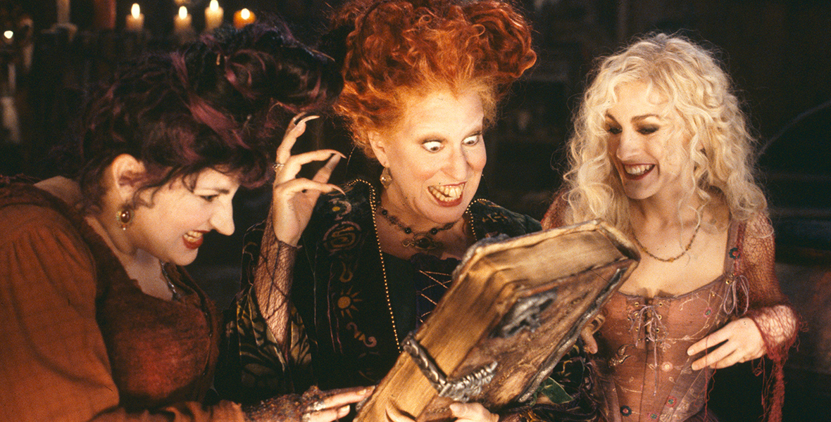 In an image from Disney’s Hocus Pocus, the Sanderson Sisters (from left to right, Kathy Najimy as Mary; Disney Legend Bette Midler as Winifred; and Sarah Jessica Parker as Sarah) are standing close together and looking down at the spell book Winifred is holding in her hand. All three are smiling, and lit candles are seen behind them.