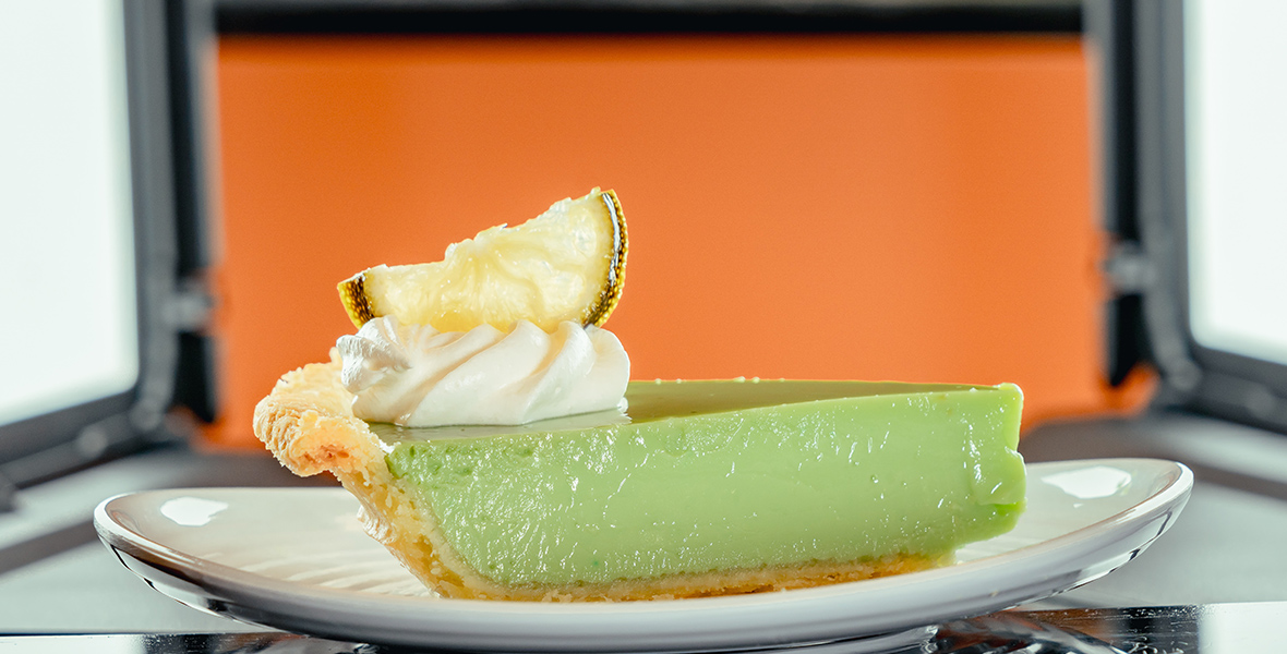 A piece of key lime pie sits on a white plate on a black table. The pie is light green with a light beige crust topped with a whipped white topping and a dried slice of lime.  
