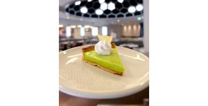 A piece of key lime pie sits on a white plate in someone’s hands. The pie is light green with a beige crust, topped with a whipped white topping and a dried slice of lemon on top. 