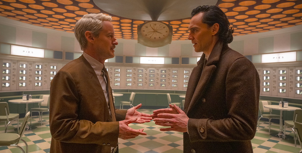In an image from the Disney+ series Loki, Mobius (Owen Wilson), left, and Loki (Tom Hiddleston), right, stand in a room filled with small diner-type tables at TVA. There’s a large clock in the background, and the ceiling—filled with orange dots—looms above and behind them. They are gesturing toward each other.