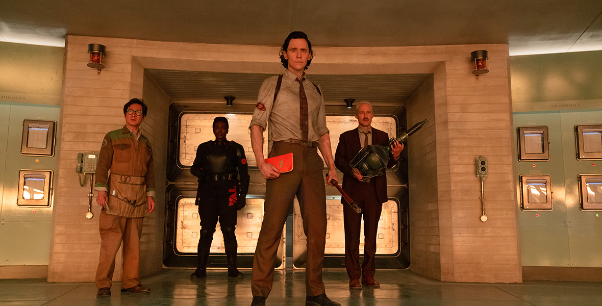 In an image from Marvel Studios’ Loki, from left to right, O.B. (Ke Huy Quan), Hunter B-15 (Wunmi Mosaku), Loki (Tom Hiddleston), and Mobius (Owen Wilson) are standing in a room looking seriously at something off-screen.