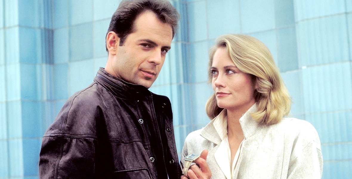 In an image from ABC’s 1980s series Moonlighting, now streaming on Hulu, David Addison (Bruce Willis), left, and Maddie Hayes (Cybill Shepherd), right, are standing in front of a building; Addison is holding a watch in one hand, and Hayes is holding jewels in both hands. He is looking straight at the camera while she is looking at him.