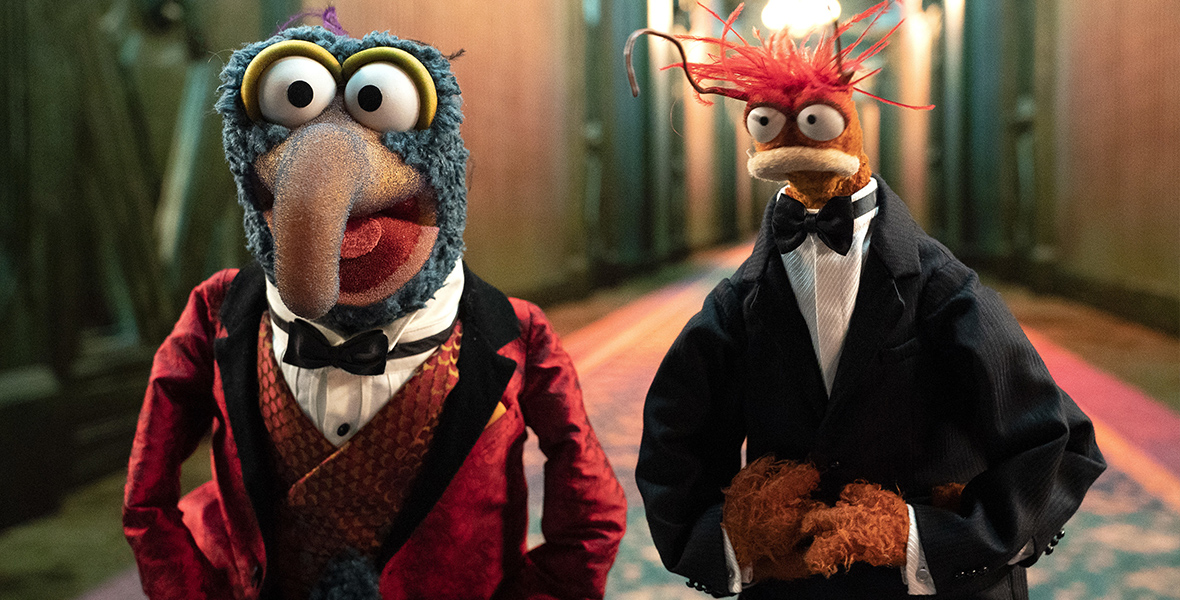 In an image from Muppets Haunted Mansion, Gonzo (voiced by Dave Goelz), left, and Pepe the King Prawn (voiced by Bill Barretta), right, are standing in one of the Haunted Mansion’s long, spooky hallways. Gonzo is wearing a red smoking jacket and white shirt and Pepe is wearing a tuxedo.