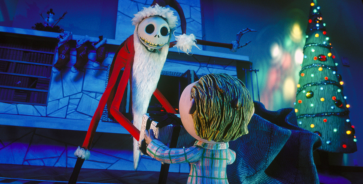 In an image from Tim Burton’s The Nightmare Before Christmas, Jack Skellington (voiced by Chris Sarandon) is standing in a living room dressed as Santa Claus, and handing a toy to a small boy whose back is toward the camera. To his right is a Christmas tree and his gift sack, and behind him is a fireplace with stockings hung above it.