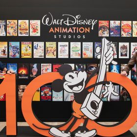 Directors Trent Correy (left) and Dan Abraham (right) pose on both sides of a giant orange 100 sculpture with a black and white Mickey Mouse in the center. Behind them are dozens of posters for Walt Disney Animation Studio's feature films and shorts.