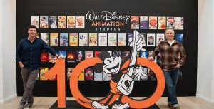 Directors Trent Correy (left) and Dan Abraham (right) pose on both sides of a giant orange 100 sculpture with a black and white Mickey Mouse in the center. Behind them are dozens of posters for Walt Disney Animation Studio's feature films and shorts.