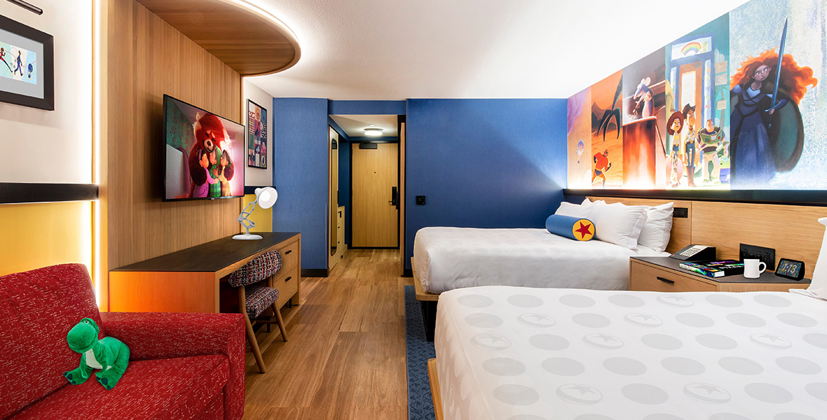 A room at the hotel features two queen-size beds with white bedspreads on the right and a built-in desk against the wall on the left, with a flat-screen television above it. Above the beds on the right is a painted montage of Pixar characters.