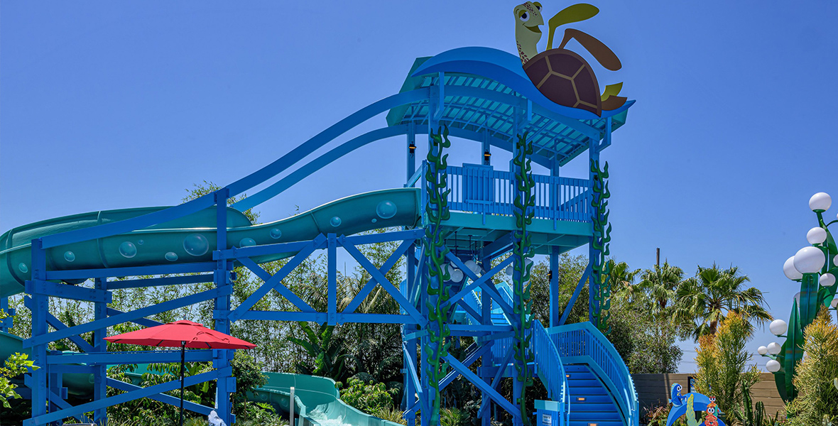 A blue tower with blue stairways leads to the top of a winding water slide that lets sliders exit into an area of fountains and pools.