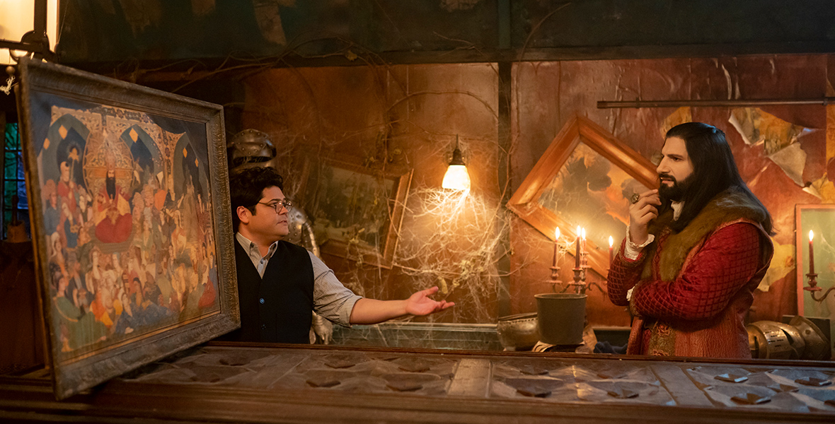 In an image from FX’s What We Do in the Shadows, Guillermo de la Cruz (Harvey Guillén), left, and Nandor the Relentless (Kayvan Novak), right, are looking at a large painting that Guillermo is holding up on a counter. The room around them is covered with cobwebs.