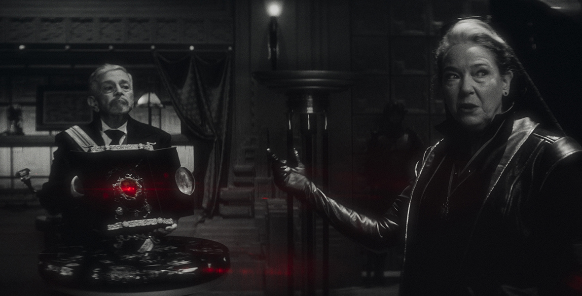 In a black-and-white image from Marvel Studios’ Werewolf by Night, Billy Swan (Al Hamacher), left, and Verussa (Harriet Sansom Harris), right, are standing in a darkened room. A suit-bedecked Swan is behind a large book displayed on a pedestal; a red light is somehow emanating from said book. Verussa, wearing a leather coat and gloves, is gesturing toward the book to people off-screen.