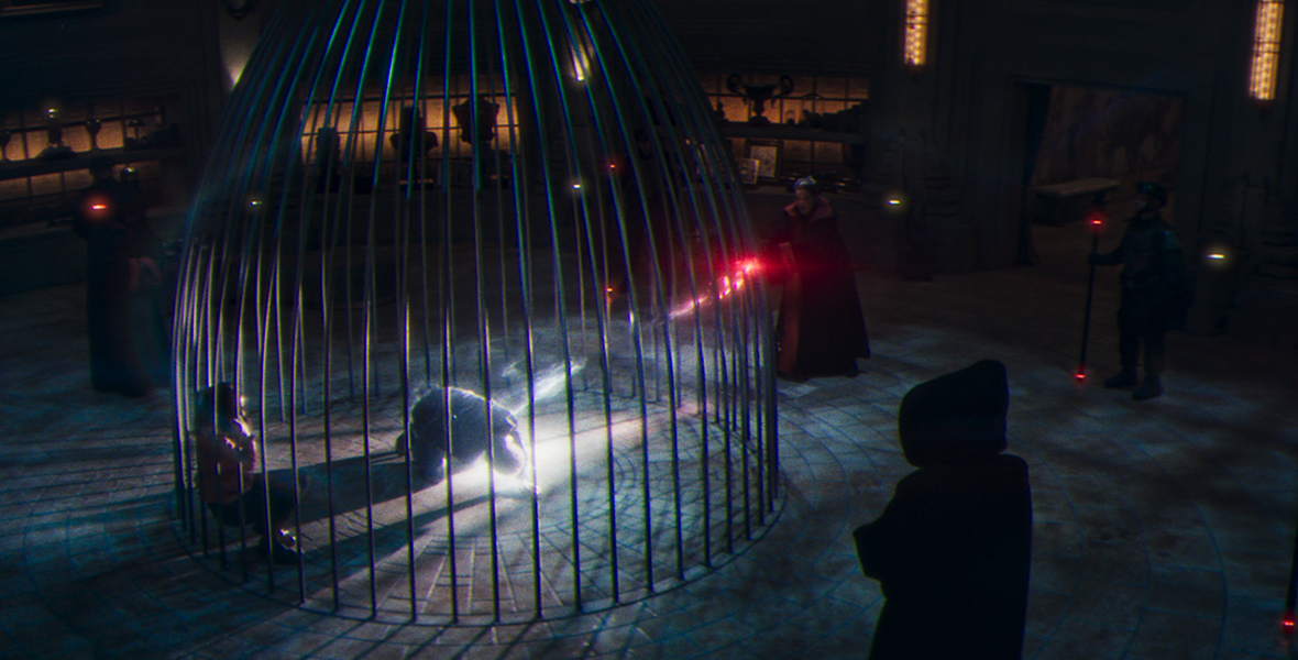 In a color image from Marvel Studios’ Werewolf by Night, two people are seen inside a huge cage that a cloaked figure, to the cage’s right, is shooting some sort of red mystical light into. There are dim lamps surrounding the perimeter of the cage. Another cloaked figure is standing below the cage, close to the camera’s vantage point.