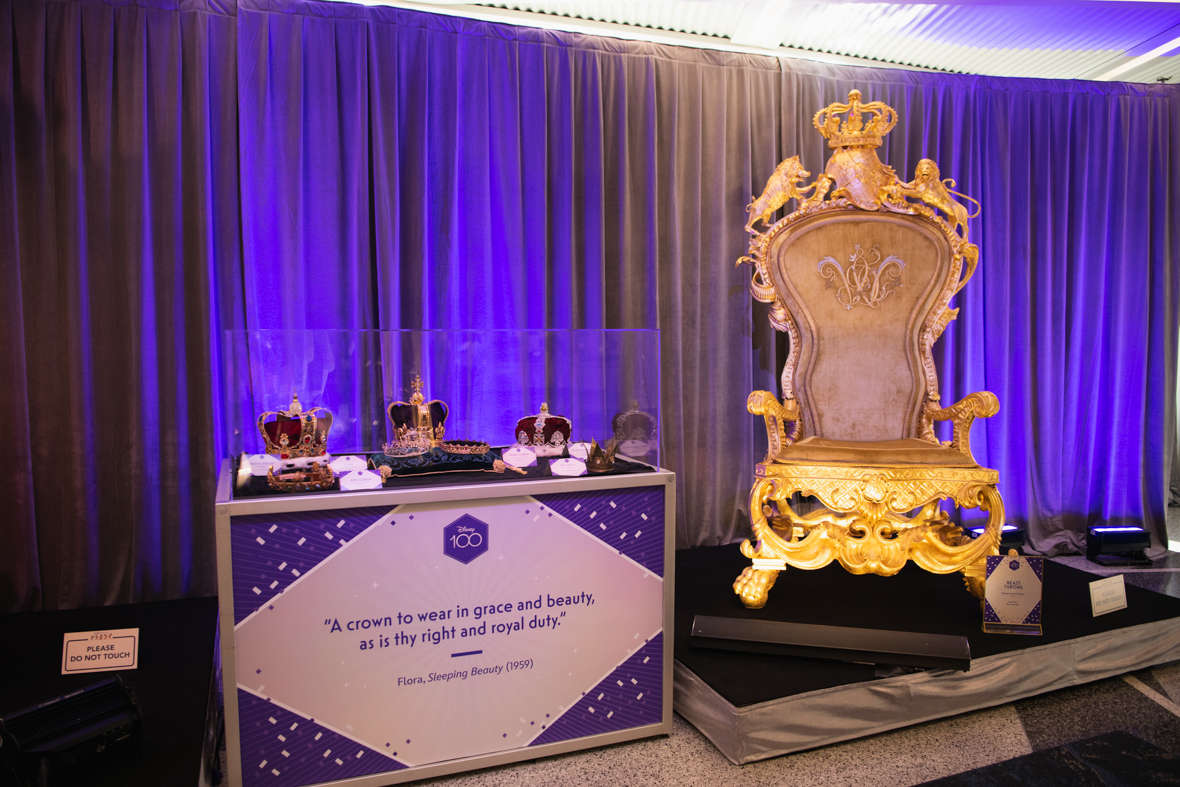 The Beast’s throne, seen in the live-action film Beauty and the Beast, is positioned on a platform at the D23 Royal Anniversary Ball’s Walt Disney Archives exhibit at the far left. At the right, a display case shows a variety of crowns and tiaras from Disney live-action films. The crown sit under glass on a table with the following written “Disney 100: ‘A crown to wear in grace and beauty, as is thy right and royal duty,’ Flora, Sleeping Beauty (1959).”