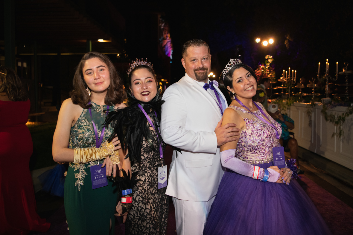 A couple pose together on the purple carpet. The man is wearing a black coat with gold detailing, black slacks, and black lipstick while holding a black and gold scepter. The woman is wearing a black dress with gold detailing, and a black sheer cape while holding a black bag with a gold apple printed on it. 