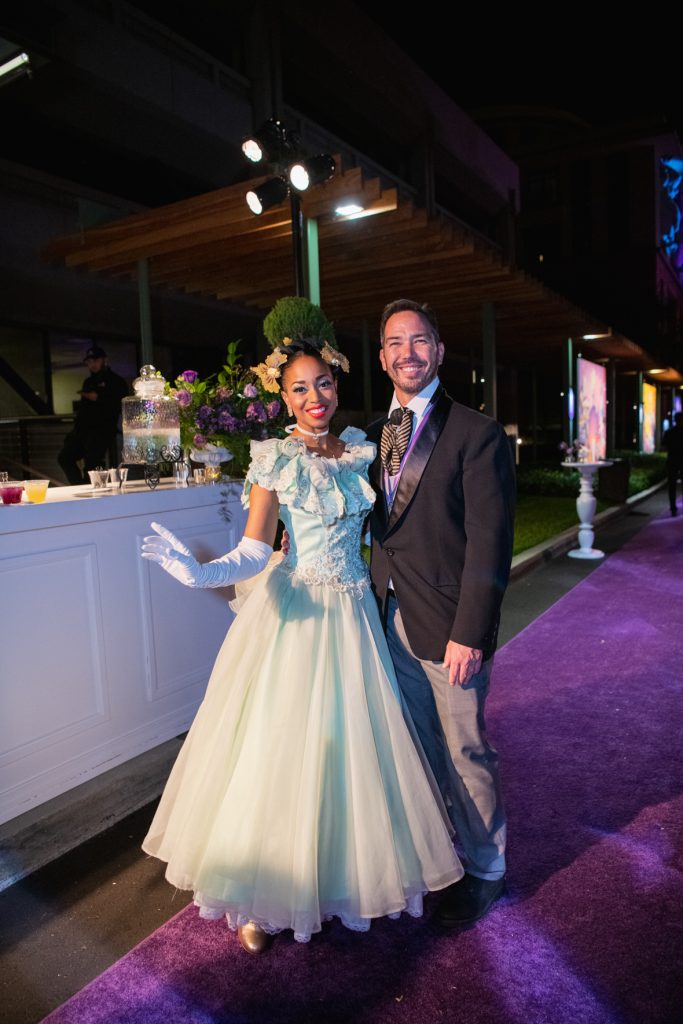 A family pose together. From left to right, a girl is wearing a green dress with gold detailing and gold gloves, next to her a girl wears a black dress with a feather shawl and tiara, next to her a man wears a white suit with a purple tie, next to him a woman wears a purple dress with purple gloves and a silver tiara. 