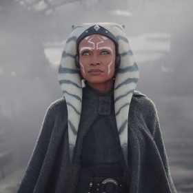 Ahsoka Tano stands in front of a spaceship with smoke coming out of it while wearing a gray cape and dress with a silver ring belt as she looks up at something in front of her.