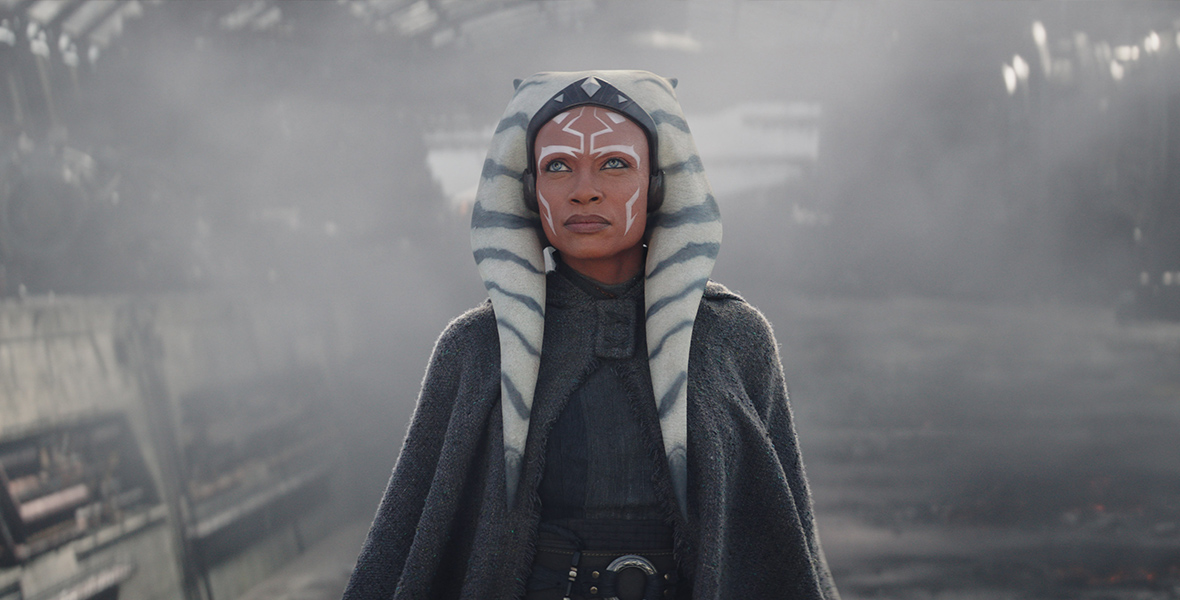 Ahsoka Tano stands in front of a spaceship with smoke coming out of it while wearing a gray cape and dress with a silver ring belt as she looks up at something in front of her.