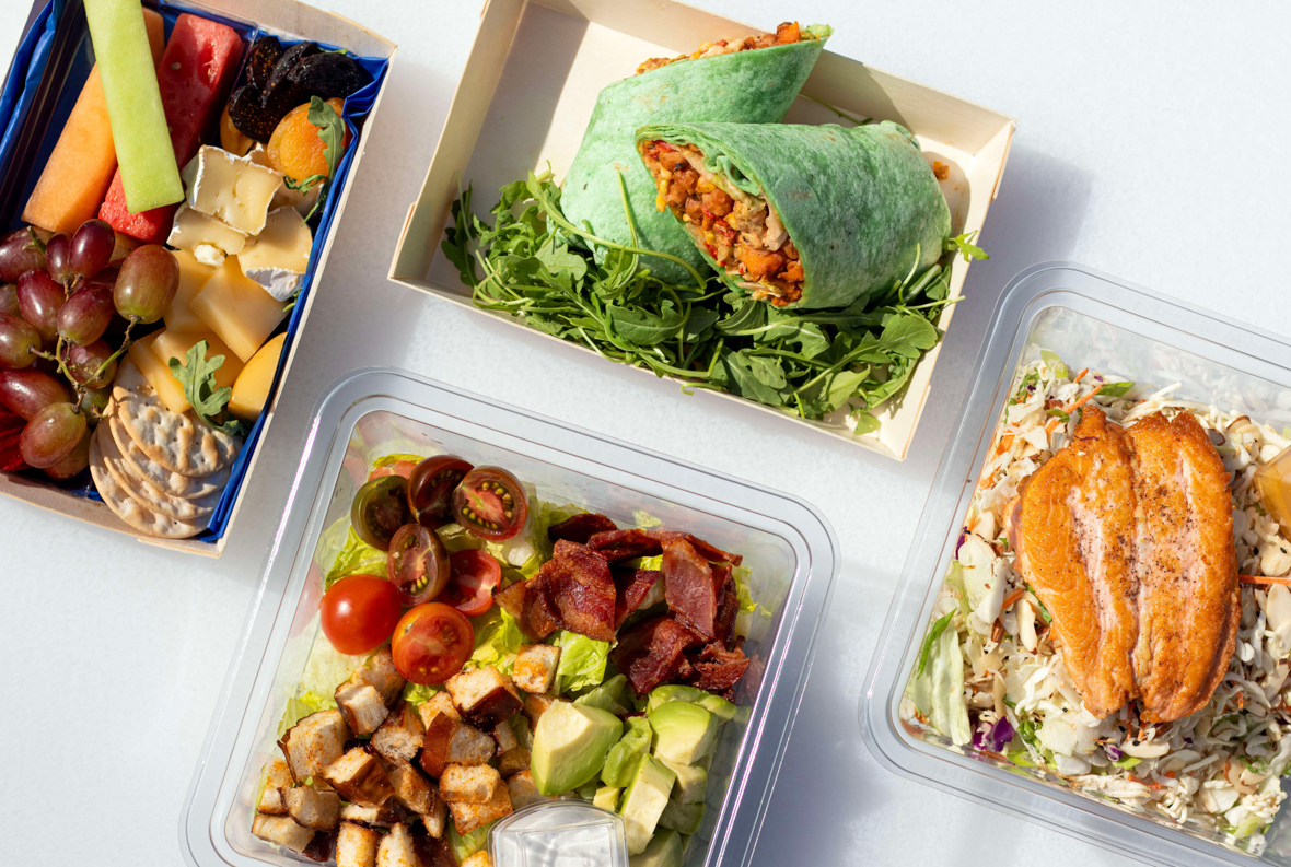 Four rectangular take-out containers display a fruit and cheese selection, a chicken wrap, a salad with protein on top, and a green salad with chopped chicken and vegetables.