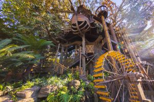 An image of the Adventureland Treehouse looking from the ground up to its peak features the working waterwheel in the foreground and the stairways behind and above it, snaking around the trunk of the banyan tree.