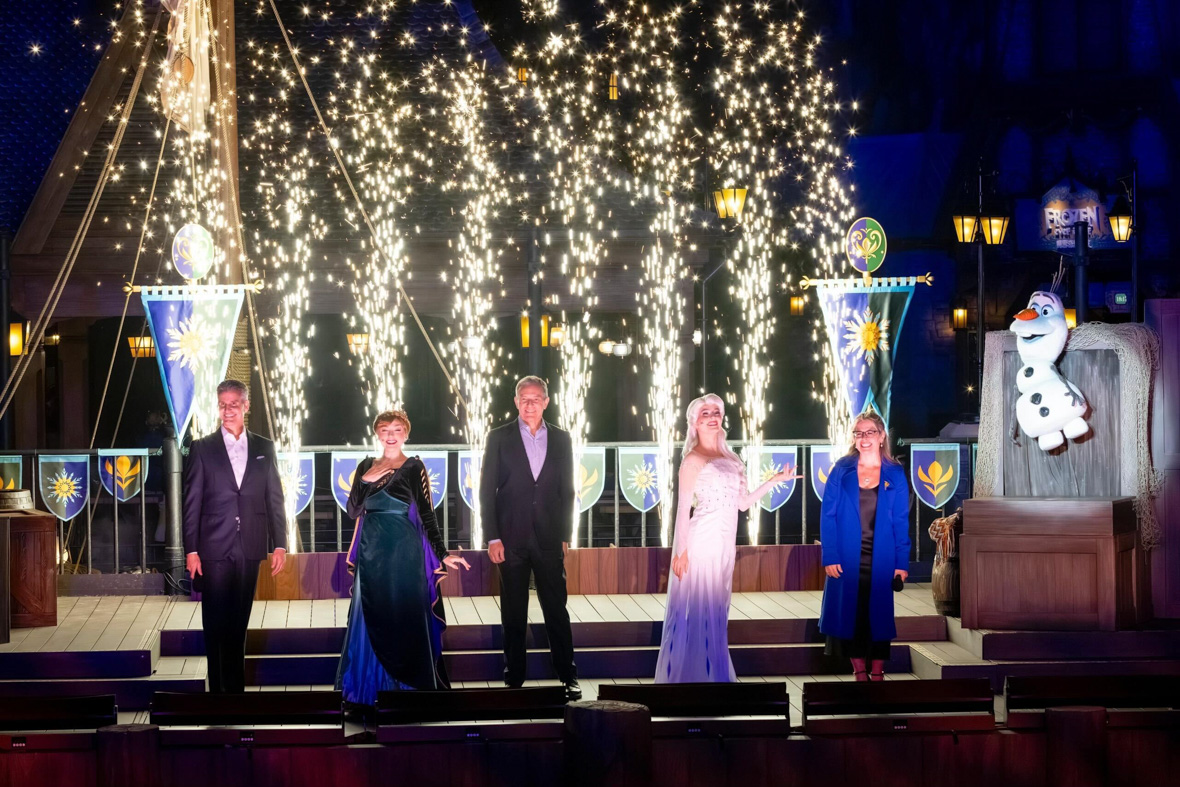 Josh D'Amaro, Anna, Bob Iger, Elsa, and Jennifer Lee smile onstage during "A Spectacular Celebration of World of Frozen" at Hong Kong Disneyland. Olaf is jumping to their left, and dazzling pyrotechnics and Arendellian flags are behind them.