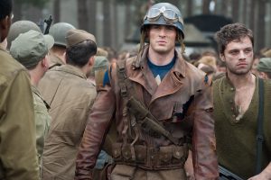Steve Rogers, played by Chris Evans, in Captain America: The First Avengeris wearing a brown jacket, blue shirt, blue army helmet, and goggles around the helmet. Bucky Barnes, played by Sebastian Stan, stands next to him wearing a green shirt, silver dog tags, and has a cut on his face. Several soldiers surround them.