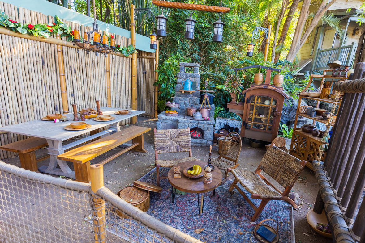 The treehouse kitchen is on the ground level. It has a picnic-style dining table on the left, and an elaborate stove in the back, next to a cabinet and some shelving. In the foreground is a seating area with a few mismatched wooden chairs arranged around a small, low table.