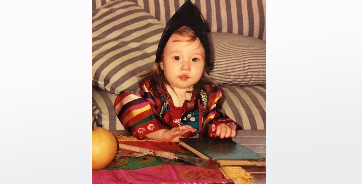 A toddler leans up against a table with objects in front of her and a striped couch behind her. She is wearing a red dress with colorful stripes and a black head dressing.