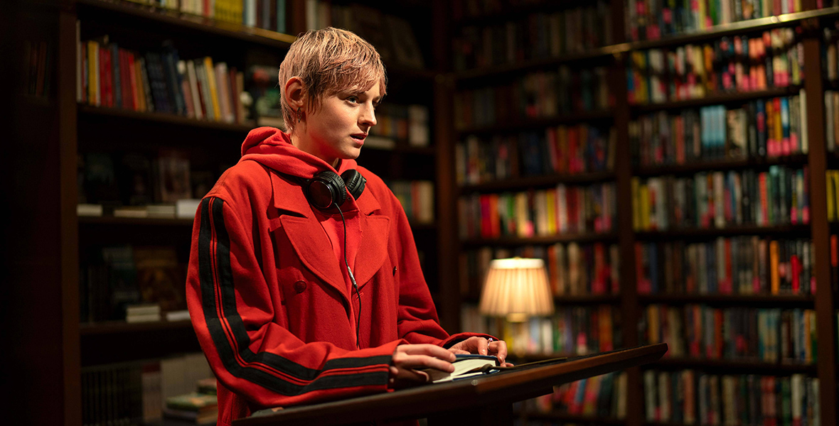 In an image from FX’s A Murder at the End of the World, Darby (Emma Corrin) is standing at a lectern in a library room, surrounded by tall bookshelves full of books—from floor to ceiling. She is wearing a red hoodie and jacket, and has headphones around her neck, and there is an open book in front of her on the lectern. 