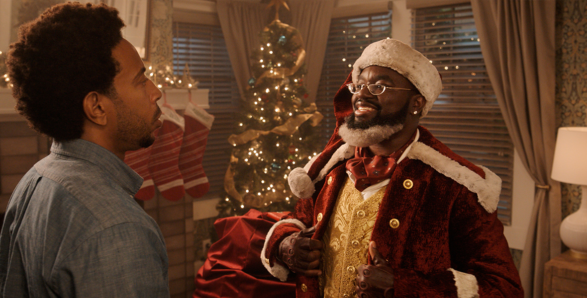 In an image from Disney+’s Dashing Through the Snow, from left to right, Eddie (Chris "Ludacris" Bridges) and Nick (Lil Rel Howery) are standing in a living room; there is a fireplace, with stockings hung on the mantel, and a Christmas tree. Nick is wearing a Santa Claus-type suit and holding a red velvet sack. He’s smiling at Eddie, who’s looking quizzically back at him.