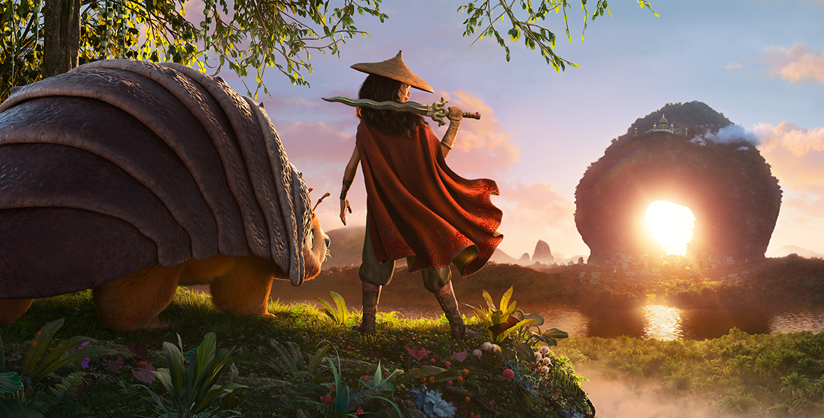 In an image from Walt Disney Animation Studios’ Raya and the Last Dragon, Raya (voiced by Kelly Marie Tran) is standing with her back to the camera, wearing a hat and holding a sword up on her right shoulder. A red cape flows out from her shoulders and down her back. Standing to her left is Tuk Tuk (voiced by Alan Tudyk), a large animal that looks like a combination of a pill bug and an armadillo. In front of the pair, to the right, is a river and a stone archway, through which the sun is shining.