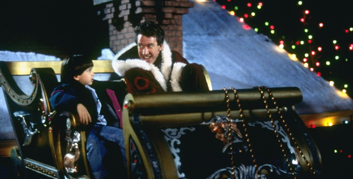 Scott Calvin (Tim Allen) and Charlie Calvin (Eric Lloyd) from The Santa Clause sit in a gold sleigh on top of a roof with a chimney in the background. Scott has a Santa suit on and is smiling at Charlie who looks up at him.
