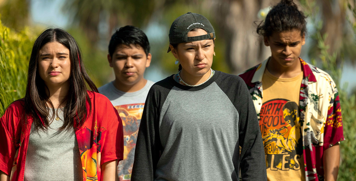 The four main characters of Reservation Dogs are all walking together. From left to right, Elora Danan, played by Devery Jacobs, is wearing a gray top with a red button down that is open. Cheese, played by Lane Factor, is wearing a gray shirt with a red and yellow print on the front. Wilhemina Jack, played by Paulina Jewel Alexis, is wearing a gray long sleeve shirt with dark grey sleeves and a black baseball cap backwards. Bear Smallhill, played by D’Pharaoh Woon-A-Tai, is wearing a yellow shirt with a yellow and red button down that is open.