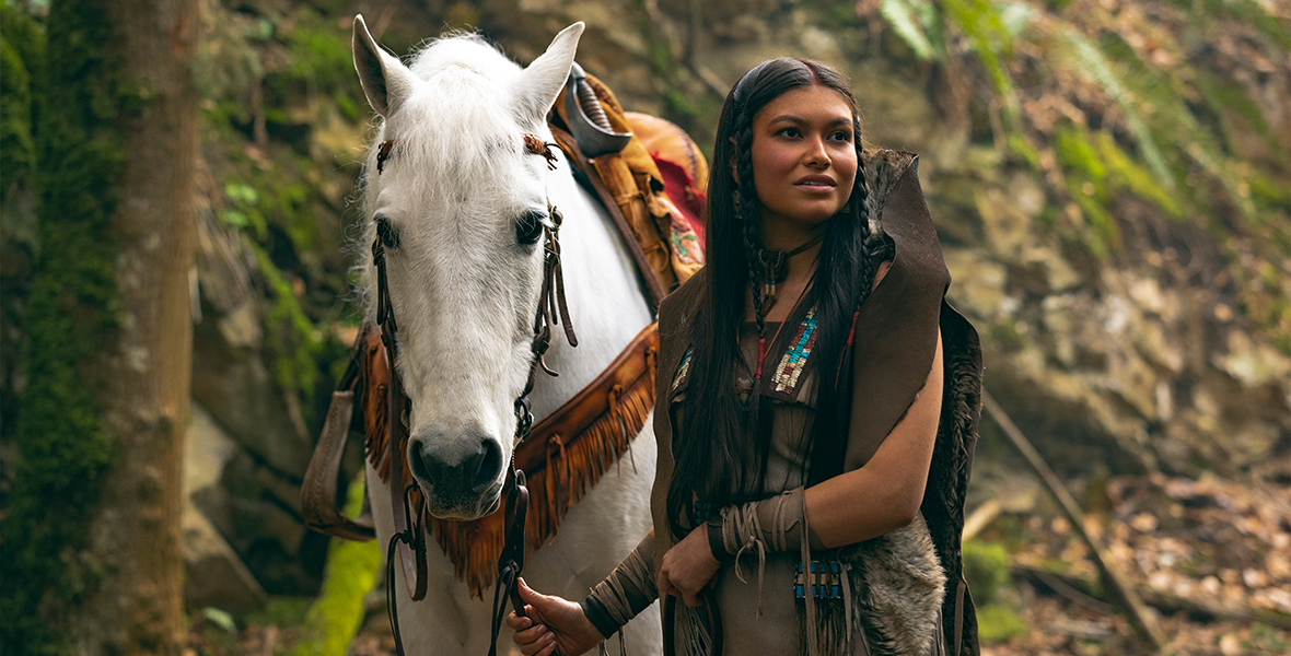 Tiger Lily from Peter Pan and Wendy, played by Alyssa Wapanatâhk, is in a brown dress with her hair styled with two braids in front. She is standing in a forest, looking off to the right while holding onto a white horse that has a light brown saddle on it. 