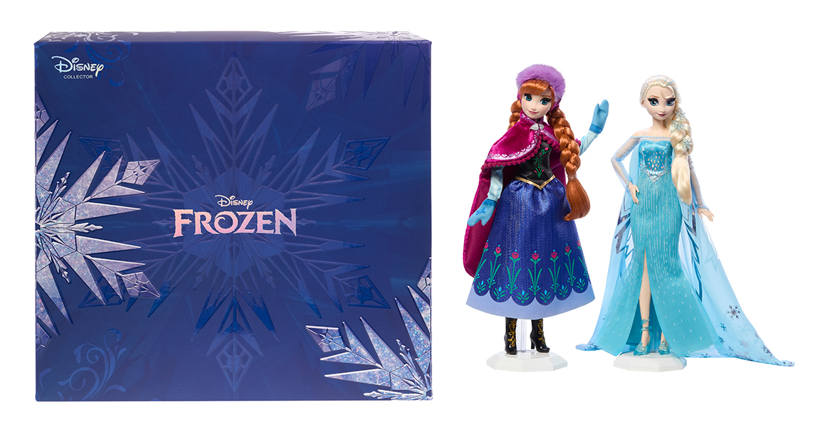 Dolls of Anna and Elsa, in their outfits from Frozen, beside their collectors box. The box is deep blue with snowflakes on the left-hand side and the lower right corner. A faint image of a snowflake sits on the center of the box, with the logo for Disney Frozen on top of it.