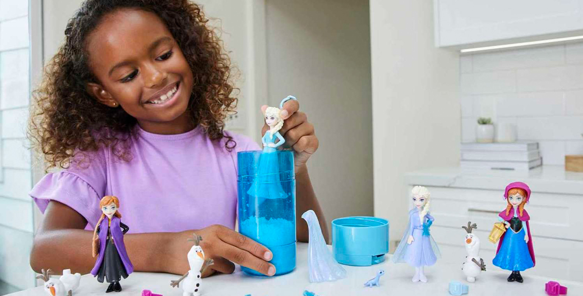 A young girl plays with small dolls, two of Anna, two of Elsa, and three of Olaf. She is removing one of the Elsas into a blue, translucent tube.