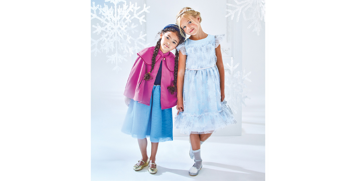Two girls stand together, holding hands and leaning on each other’s shoulders. One girl is wearing a cape and dress inspired by Anna, while the other girl is wearing a light blue dress inspired by Elsa.